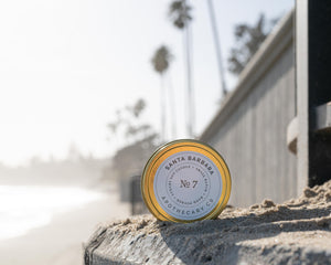 Small batch soy candle in gold travel tin on sandy sea wall with beach palm trees